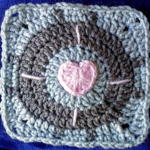 May include: A crocheted square with a pink heart in the center. The square is made with blue and gray yarn and has a white border.