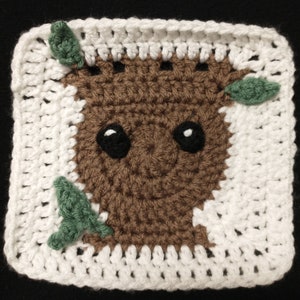 May include: A crocheted square featuring a brown and green character with a face, resembling a tree. The character is surrounded by a white crocheted border.