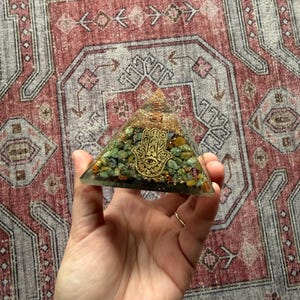 May include: A clear resin pyramid filled with green and yellow stones, topped with gold-colored wire and a gold Hamsa hand symbol. The pyramid is held in a hand, with a patterned rug in the background. The pyramid is a decorative item.