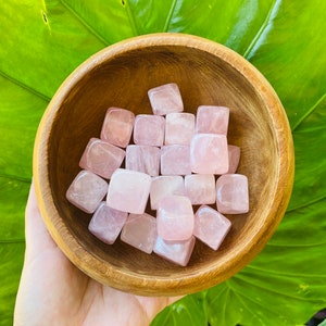 May include: A wooden bowl filled with pink rose quartz tumbled stones. The bowl is being held by a hand.