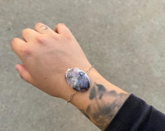 Op de afbeelding: Een armband met een ovale steen in paarse, grijze en witte tinten, bevestigd aan een fijne zilveren ketting. De armband is een sieraad. De steen is ongeveer 3 cm lang.