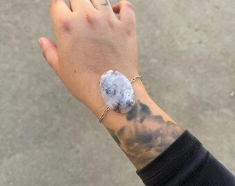 Op de afbeelding: Een zilveren armband met een ovale, wit en grijze steen. De steen is verbonden met een fijne ketting. De armband wordt gedragen om een pols met een zwarte mouw.