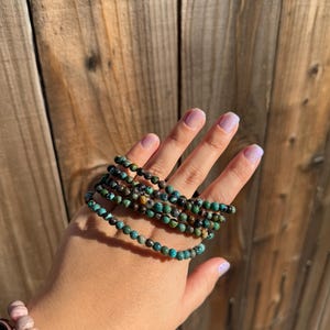 May include: A set of five turquoise and brown beaded bracelets stacked on a person's wrist. The bracelets are made of small, round beads.