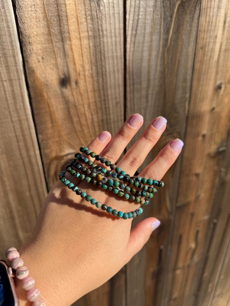 May include: A set of five turquoise and brown beaded bracelets stacked on a person's wrist. The bracelets are made of small, round beads.