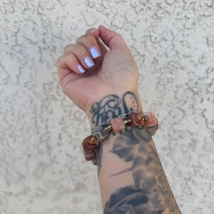 K&ouml;nnte beinhalten: Ein Armband aus rosa und braunen Steinperlen mit goldenen Akzenten. Das Armband wird am Handgelenk getragen.