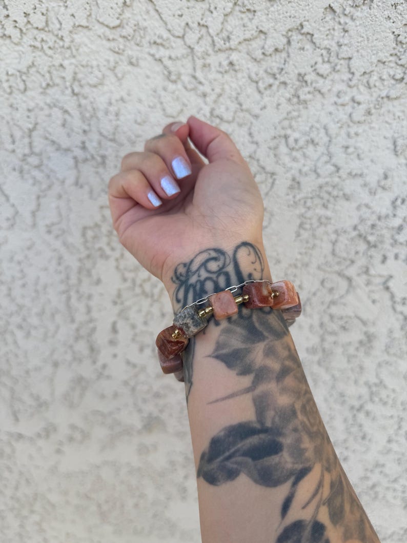 K&ouml;nnte beinhalten: Ein Armband aus rosa und braunen Steinperlen mit goldenen Akzenten. Das Armband wird am Handgelenk getragen.