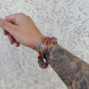 K&ouml;nnte beinhalten: Ein Armband mit goldenen Kettengliedern und quadratischen, mehrfarbigen Steinen. Die Steine sind eine Mischung aus Rosa, Orange und Wei&szlig;.