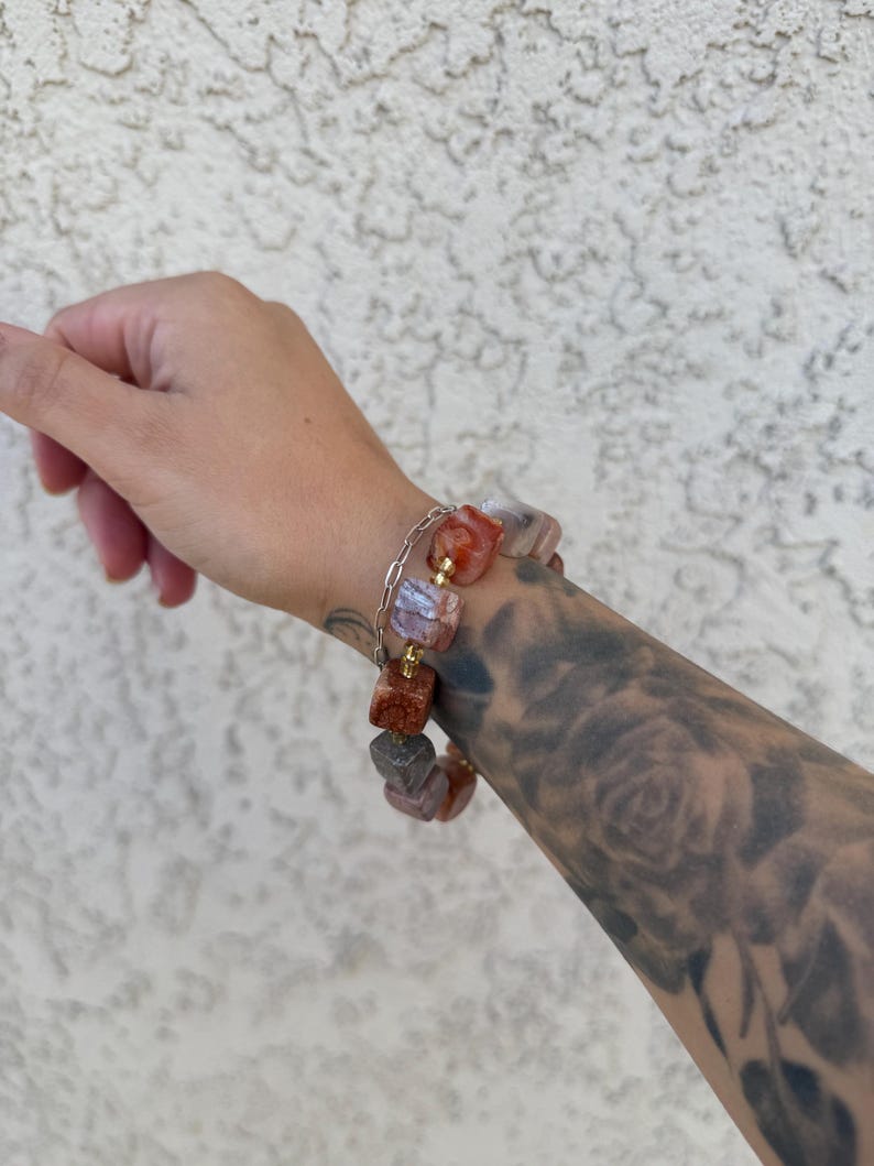 K&ouml;nnte beinhalten: Ein Armband mit goldenen Kettengliedern und quadratischen, mehrfarbigen Steinen. Die Steine sind eine Mischung aus Rosa, Orange und Wei&szlig;.