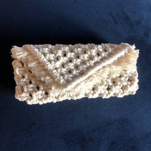 以下が含まれることがあります： フリンジのディテールが付いた白いクロシェのクラッチバッグ。バッグは厚みのある質感のあるコードで作られており、シンプルで幾何学的なデザインです。