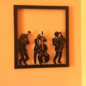 Può includere: Arte da parete in metallo nero con la silhouette di tre musicisti che suonano strumenti. I musicisti sono un trombettista, un bassista e un sassofonista. L'arte è incorniciata in una cornice nera.