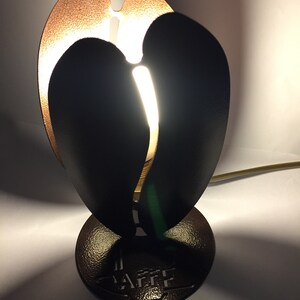 Op de afbeelding: Een decoratieve tafellamp met een uniek ontwerp. De lamp heeft een zwarte, hartvormige kap met een koperkleurig interieur. De basis is een getextureerde, bronskleurige cirkel. Het woord "CAFFE" is in de basis geëmbosseerd.