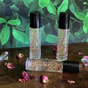 May include: Three clear glass roller bottles with black caps filled with a liquid containing gold glitter. The bottles are on a wooden surface with dried rose petals.