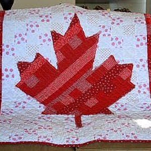 May include: A red, white, and pink quilt with a large red maple leaf in the center. The quilt is made of patchwork squares and is perfect for a Canadian themed home.
