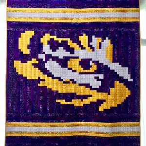 Puede incluir: Una colcha morada y dorada con un diseño pixelado. El diseño es una imagen estilizada de la cabeza de un tigre.