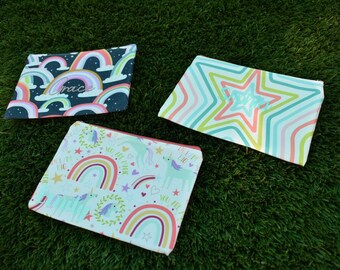 May include: Three colorful zippered pouches with whimsical designs. One features a rainbow and the name "Grace", another has a star pattern, and the third showcases unicorns, rainbows, and text. The pouches are set against a green background.