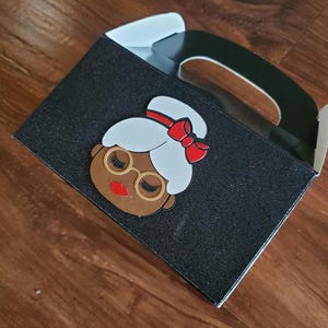 May include: Black favour box with a glitter finish. The box has a handle and features a cartoon image of a woman with a red bow in her hair.