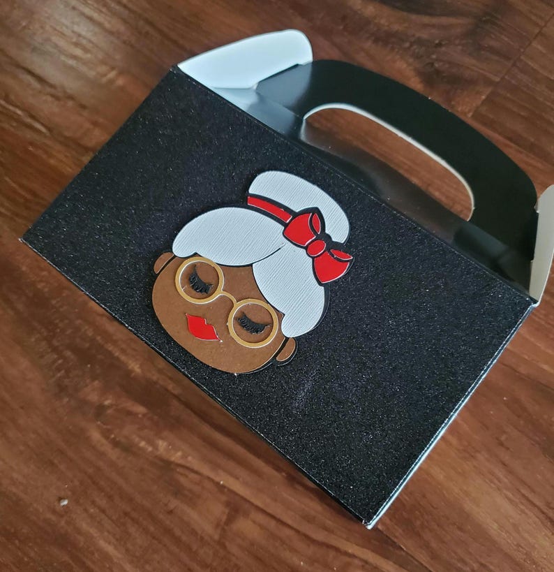 May include: Black favour box with a glitter finish. The box has a handle and features a cartoon image of a woman with a red bow in her hair.