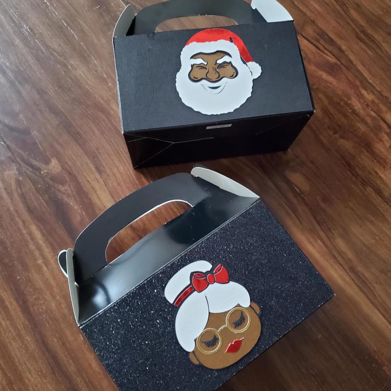 May include: Two black favour boxes with handles. One box has a glitter finish and features a cartoon image of a woman with a red bow in her hair. The other box features a cartoon image of a man with a red hat. Both images are of people with brown skin.