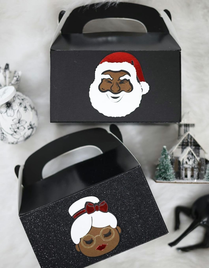 May include: Two black favour boxes with glitter. One box features a black Santa Claus with a red hat and white beard. The other box features a black woman with a red bow in her hair and glasses.