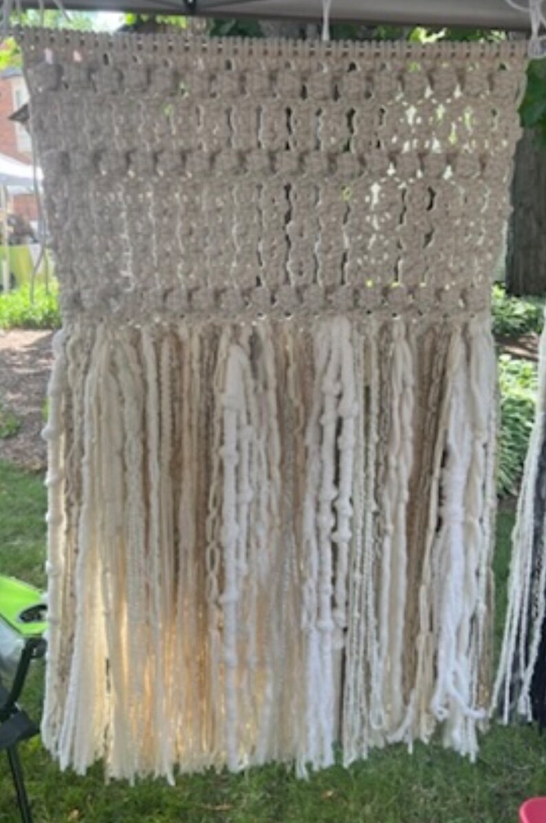 Op de afbeelding: Een beige macram&eacute; wandhanger met een geweven bovenkant en lange, witte en beige garen franjes.