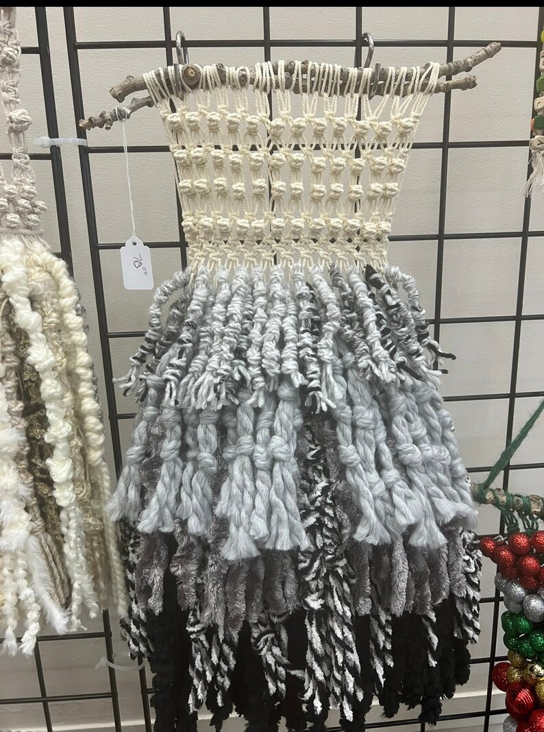 May include: A white and grey macrame wall hanging with black and white fringe. The hanging is made with natural materials and has a rustic, bohemian style. It is 198 cm long.