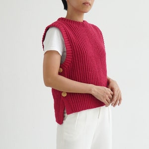 May include: A red knitted sleeveless sweater vest with wooden buttons on the side. The vest is worn over a white t-shirt and white pants.