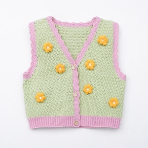 以下が含まれることがあります： ピンクの縁取りと黄色の花の装飾が施された緑色のクロシェ編みベスト。ベストはボタン留めです。