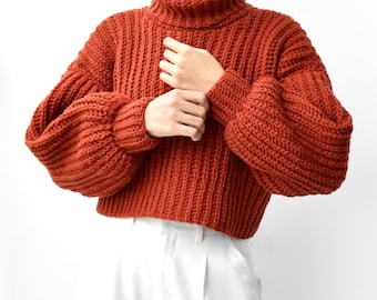 May include: A rust-colored, chunky knit sweater featuring a high turtleneck collar and long, voluminous sleeves with ribbed cuffs. The garment has a thick, vertical ribbed texture throughout and a cropped hemline, paired with white trousers.