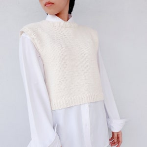 Peut inclure: Un gilet sans manches en tricot blanc avec un col rond. Le gilet est superposé à une chemise boutonnée blanche.