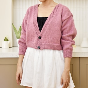 May include: A pink cropped cardigan with a V-neck and button closure. The cardigan is made of a soft, knit fabric.