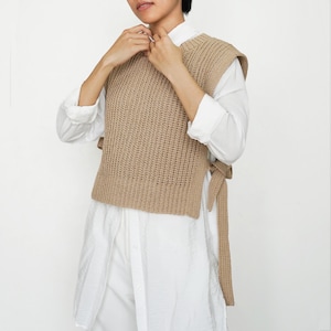 Può includere: Un gilet in maglia senza maniche color beige con una trama a coste e lati aperti con lunghi lacci in maglia abbinati. Il gilet è indossato sopra una camicia bianca a maniche lunghe con colletto classico e polsini arrotolati.