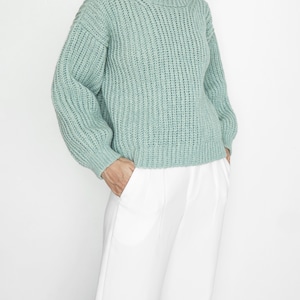 May include: A sage green long-sleeve sweater featuring a chunky ribbed knit texture, a rounded crew neckline, and dropped shoulders, paired with crisp white trousers that have a straight-leg cut and a clean, tailored appearance.