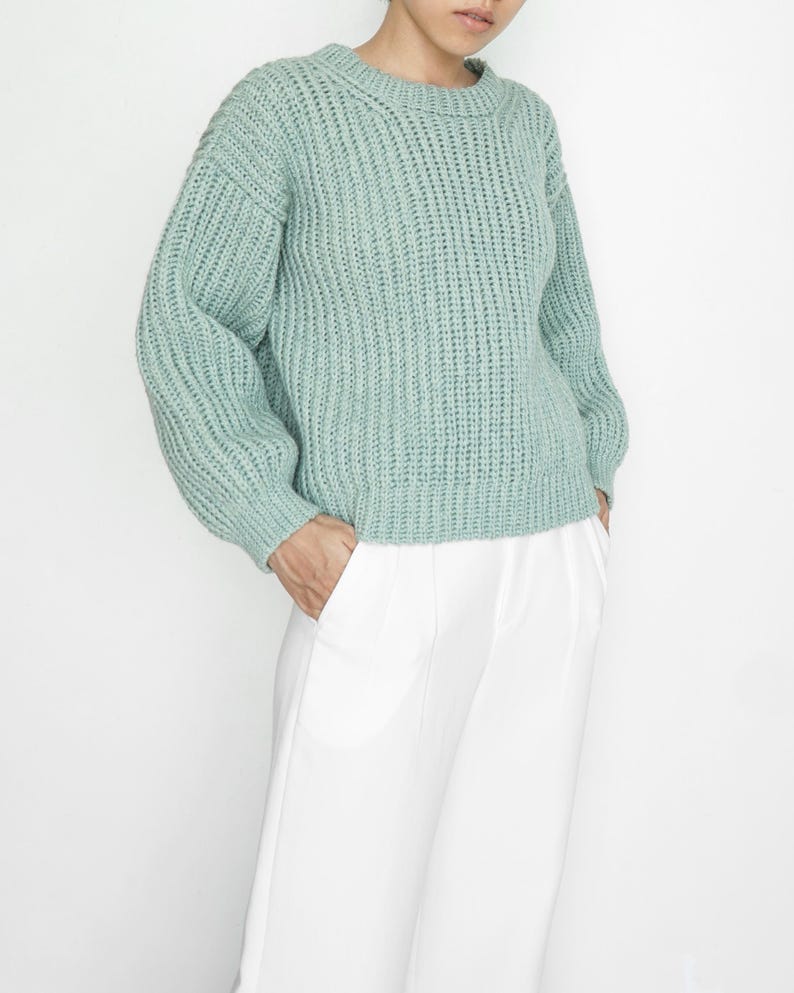 May include: A sage green long-sleeve sweater featuring a chunky ribbed knit texture, a rounded crew neckline, and dropped shoulders, paired with crisp white trousers that have a straight-leg cut and a clean, tailored appearance.