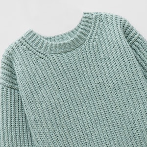 May include: A light sage green long-sleeve sweater featuring a chunky knit texture, a ribbed crew neckline, and ribbed cuffs. The garment is constructed with a thick, vertical knit pattern throughout the body and sleeves, providing a consistent appearance.