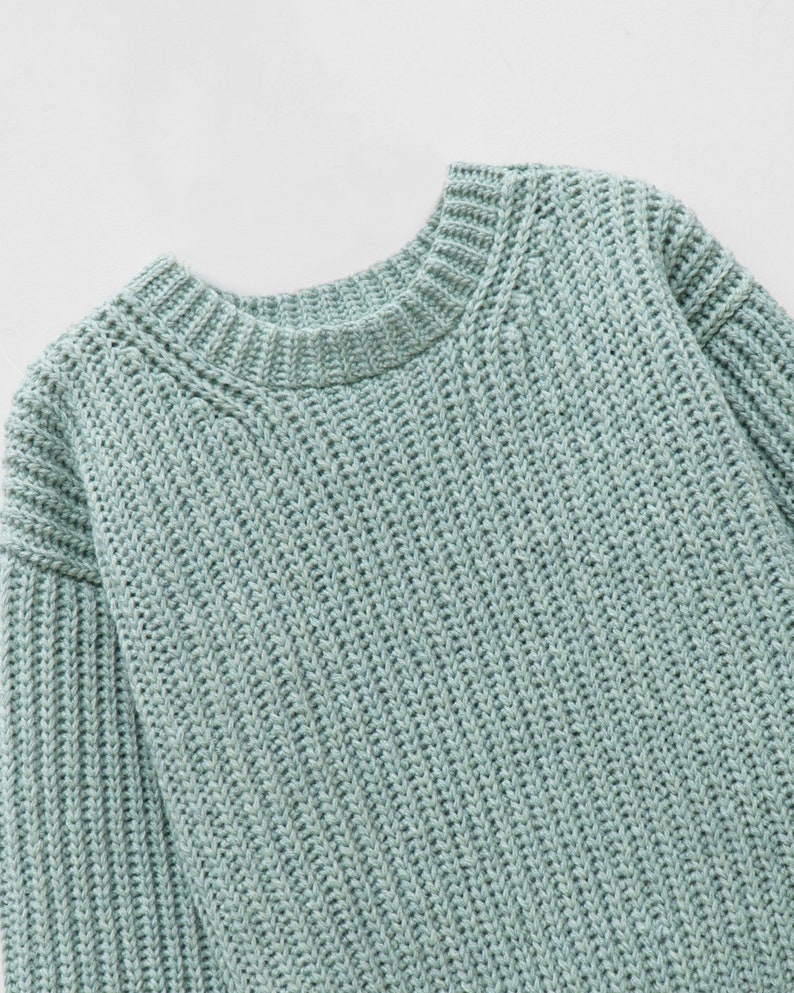 May include: A light sage green long-sleeve sweater featuring a chunky knit texture, a ribbed crew neckline, and ribbed cuffs. The garment is constructed with a thick, vertical knit pattern throughout the body and sleeves, providing a consistent appearance.