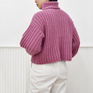 May include: A pink, chunky knit turtleneck sweater with a cropped hem. The sweater is made with a ribbed stitch and has a relaxed fit.