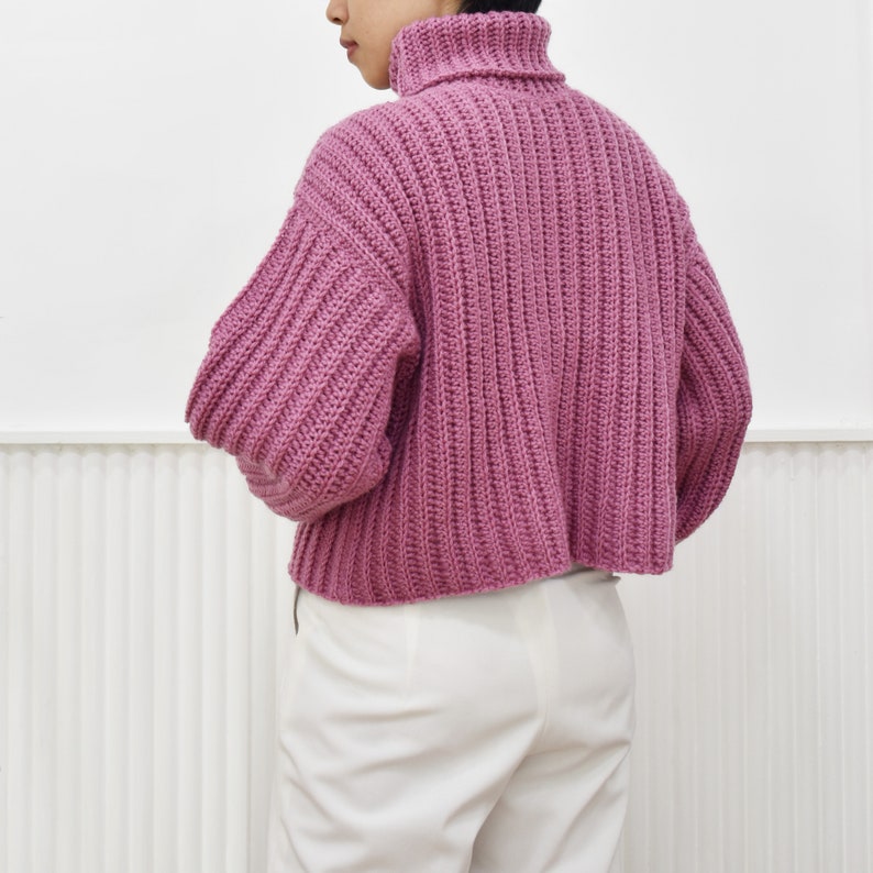 May include: A pink, chunky knit turtleneck sweater with a cropped hem. The sweater is made with a ribbed stitch and has a relaxed fit.