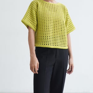 May include: A lime green, open-weave crochet top with short sleeves. The top has a round neckline and a loose fit. The model is wearing black pants. The top is a handmade garment, perfect for warm weather.