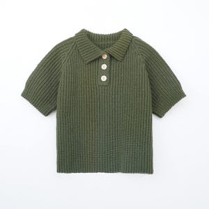 May include: Olive green knitted polo shirt with short sleeves and a collared neckline. The shirt features three wooden buttons down the front. The knitwear has a textured appearance, and the garment is displayed on a white background.