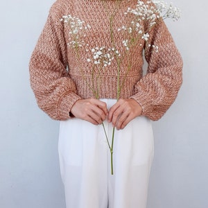 May include: A brown and white knit sweater with a cropped length and long sleeves. The sweater is worn by a person holding a bouquet of white baby's breath flowers.