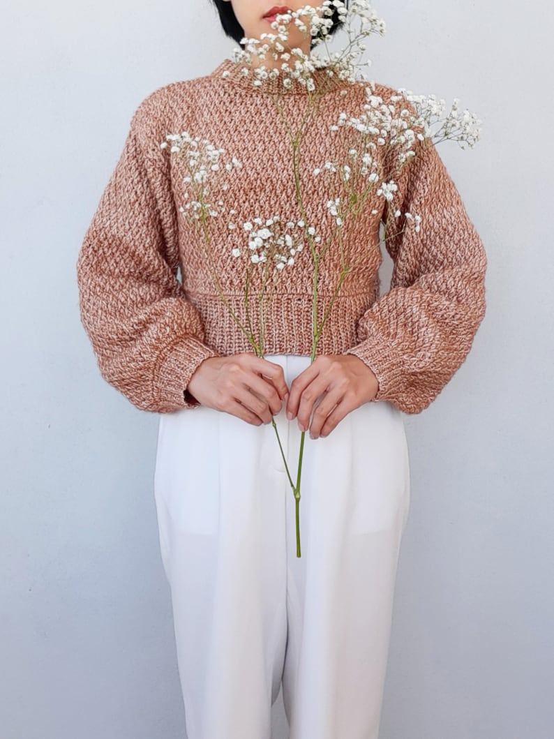 May include: A brown and white knit sweater with a cropped length and long sleeves. The sweater is worn by a person holding a bouquet of white baby's breath flowers.