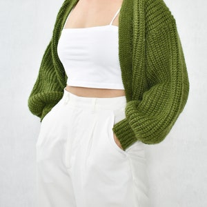 May include: A green knitted cardigan with long sleeves and a V-neck. The cardigan is worn over a white vest top and white trousers.