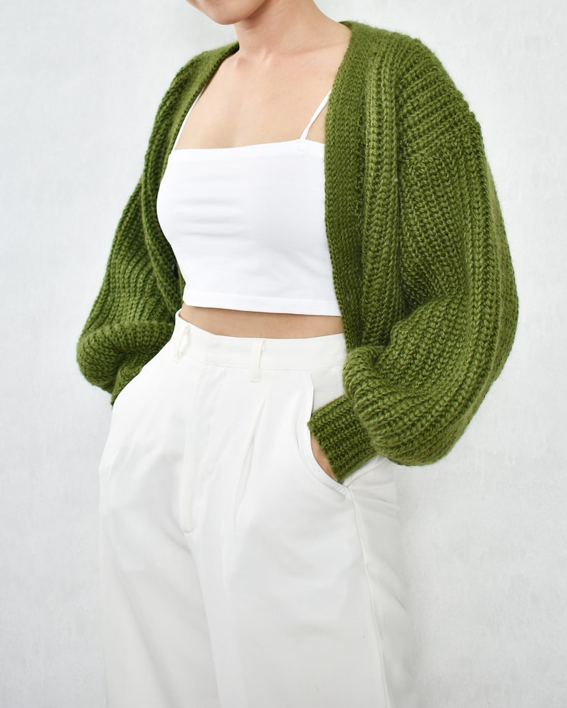 May include: A green knitted cardigan with long sleeves and a V-neck. The cardigan is worn over a white vest top and white trousers.