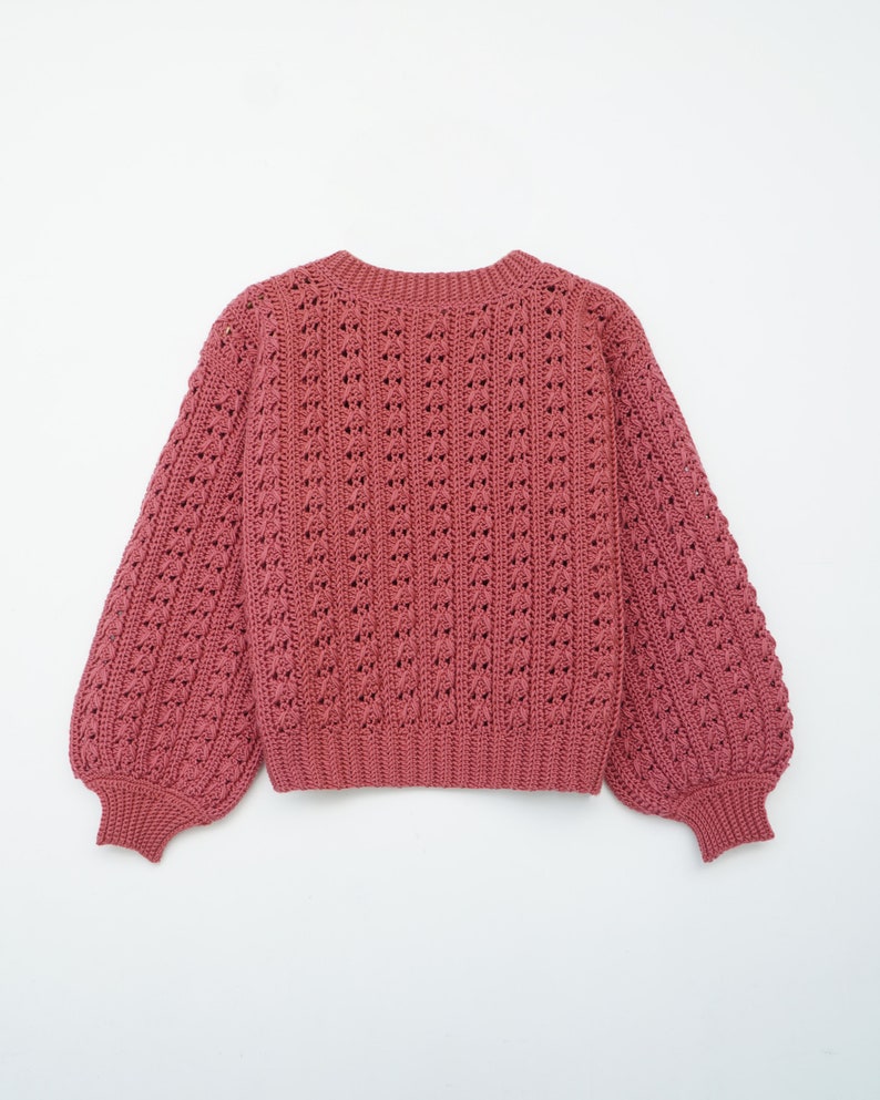 May include: A pink crocheted sweater with a loose fit and long sleeves. The sweater has a delicate openwork pattern.