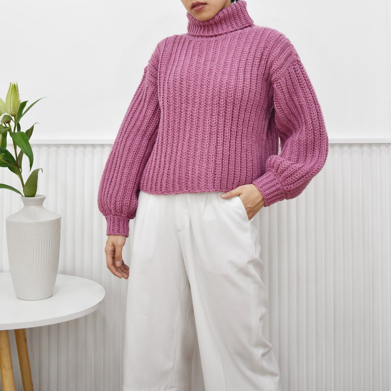 May include: A pink knitted turtleneck sweater with long sleeves and a cropped fit. The sweater has a ribbed texture and a relaxed fit.