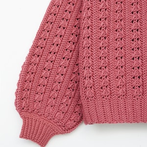 May include: A close-up of a pink crocheted sweater with a textured pattern. The sweater has long sleeves and a loose fit.