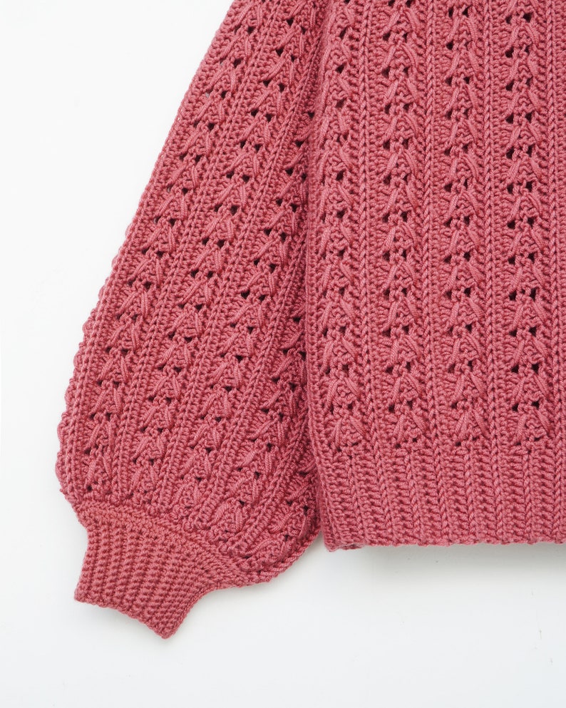 May include: A close-up of a pink crocheted sweater with a textured pattern. The sweater has long sleeves and a loose fit.
