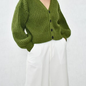 May include: A green knitted cardigan with a v-neck and button closure. The cardigan has a relaxed fit and long sleeves.