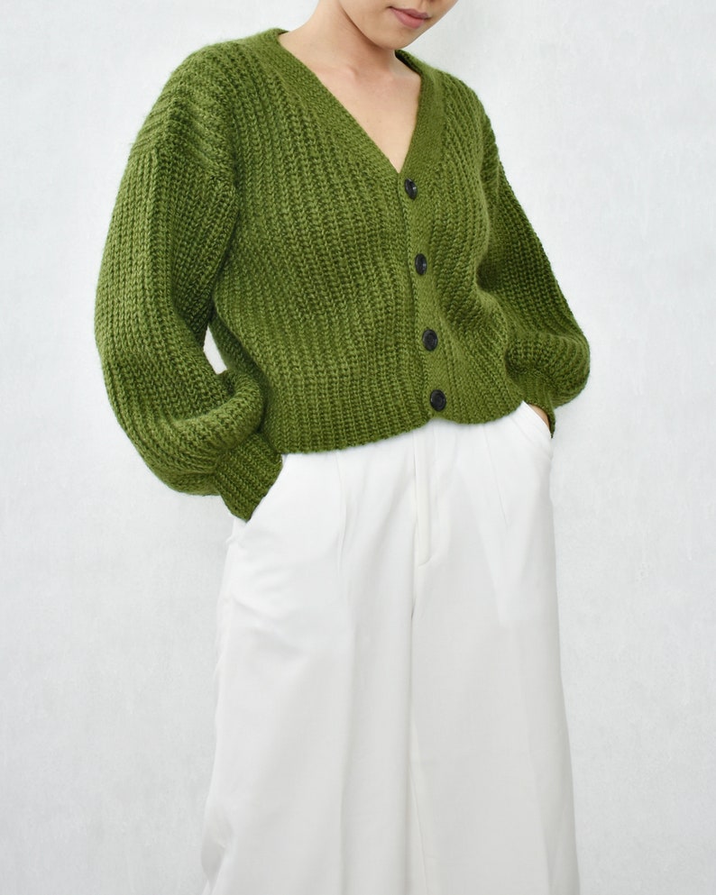 May include: A green knitted cardigan with a v-neck and button closure. The cardigan has a relaxed fit and long sleeves.