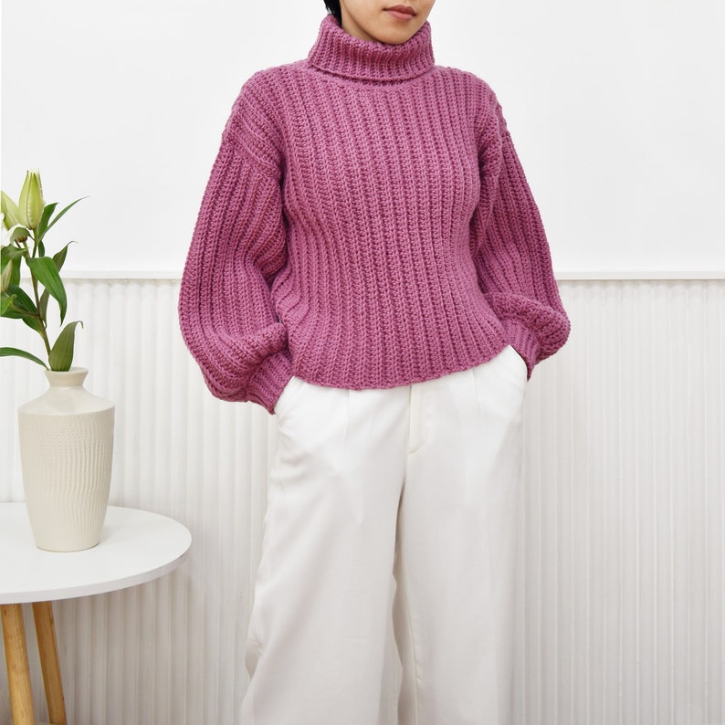 May include: A pink, chunky knit turtleneck sweater with long sleeves and a slightly cropped fit.